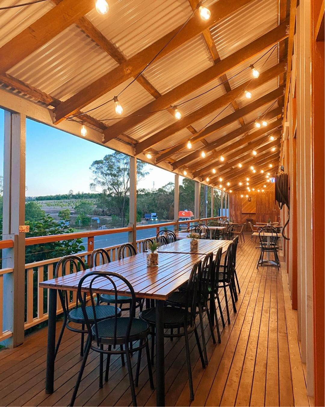 A rustic outdoor deck at dusk illuminated with festoon lights, featuring long tables and matching chairs.
