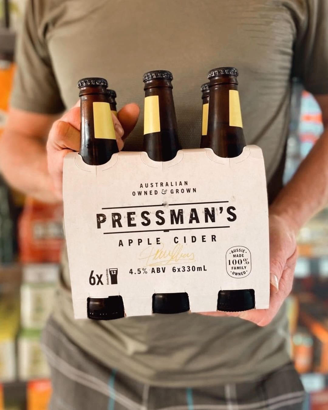 A man holding a six pack of Pressman's Apple Cider. You can see the bottle shop fridges blurred out in the background.